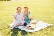 Two young beautiful smiling hipster female in summer sundress and hats. Carefree women making picnic outside. Positive models sitting on plaid on grass, drinking champagne,  eating fruits