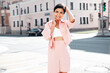 Young beautiful smiling hipster woman in trendy summer pink costume clothes. Carefree female posing in the street at sunny day. Positive model outdoors at sunset. Cheerful and happy