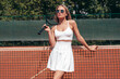Young beautiful woman wearing fashionable white dress skirt and tank top. Hot model posing on tennis court at summer sunny day. Sexy female stands near net