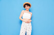Young beautiful smiling female in trendy summer clothes. Carefree woman posing near blue wall in studio. Positive model having fun. Cheerful and happy. With curly short hairstyle, in hat