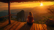© john - Picture from the back of a woman sitting on wooden porch extending into a high mountain cliff. The sun is setting on the mountain and there is a beautiful warm orange light. The traveling background.