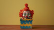 Orange Haired Clown Toy Popping out of a Blue and Yellow Box on Mustard Background