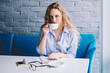 Portrait of charming caucasian hipster girl sitting at cafeteria and drinking caffeine beverage, beautiful woman holding cup with hot tea and looking at camera in break of studying in cafe interior