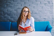 Portrait of attractive hipster girl holding literature in hands and enjoying time for hobby in cafeteria, beautiful woman in fashionable spectacles looking at camera during reading best seller