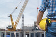 A male construction worker holds a yellow helmet, standing in front of a busy construction site with cranes and unfinished buildings.