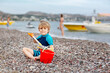 Adorable little blond kid boy building pebble stone castle on beach. Funny child playing with bucket and shovel. Vacations, summer, travel concept. Toddler enjoying summer vacations on sea.