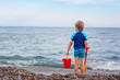 Adorable little blond kid boy standing on lonely ocean beach. Child playing with bucket and shovel and looking on horizon. Vacations, summer, travel concept. Summer family vacations on sea.