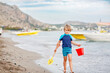 Adorable little blond kid boy walking on ocean beach. Child playing with bucket and shovel and looking on horizon. Vacations, summer, travel concept. Summer family vacations on Mediterranean Sea