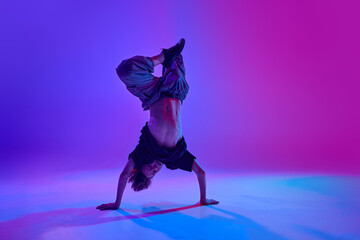 Stylish dressed young dancer, guy performing headstand in motion in mixed neon light against vibrant gradient background. Concept of sport and hobby, music, fashion and art, movement. Ad