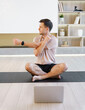 A man sits cross-legged on a yoga mat in a well-lit room, stretching his arms while using a laptop for guidance.