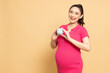 Happy Asian pregnant woman standing and holding piggy bank isolated on brown background, Saving in bank and financial with pregnancy concept