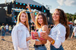 © artiemedvedev - Group of happy females cheers with cocktails at a lively beach music festival. Smiling friends enjoying party, summer vibe, coastline backdrop. Girls toast drinks, celebrate, dance to live band.