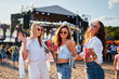 © artiemedvedev - Girls enjoying summer music festival on beach, holding drinks, dancing in sunshine. Group of young women celebrate, having fun at outdoor concert, vibrant fest scene with crowd, stage in background.