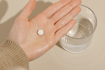 Wall Mural - Female hand holding a white pill against a glass of water on a beige isolated background. The concept of taking medicines, dietary supplements, health care.