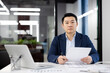 Focused Asian businessman sitting at desk in a well-lit office, reviewing documents with serious expression, displaying professionalism and determination.