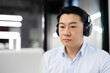 A professional Asian male in business attire focuses intently while wearing headphones. He appears engaged and serious, sitting in a modern office.