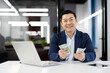 A cheerful Asian businessman counting US dollars at his desk in a well-lit modern office. He exudes success and financial security with a joyful expression.