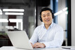 Cheerful Asian businessman with headphones engaged in online communication while using a laptop in a modern office setting.