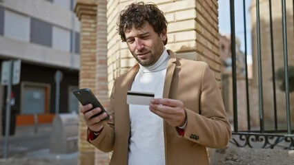 Sticker - A young man using a smartphone and holding a credit card on an urban street.