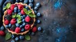   A pie filled with berries - blueberries and raspberries - rests on a black table against a blue backdrop Berries spill over the edge, mint leaves adorn