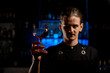 Bartender holds a high-stemmed glass filled with a cocktail in his hand at the level of his face and shakes it so
