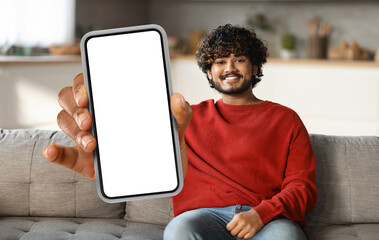 Poster - Eastern man is seated on a couch, holding a smart phone in his hand. He appears focused on the devices screen, possibly browsing or messaging.