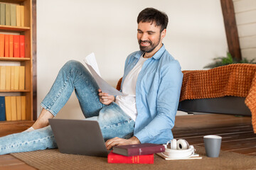 Poster - A cheerful man in casual attire sits on the floor with his laptop open and paperwork in hand, sitting area features a coffee cup, headphones, and books, suggesting a relaxing work-from-home setup