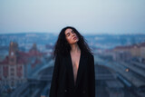 Fototapeta Zwierzęta - Young attractive brunette in a black jacket stands on the roof with closed eyes against the background of the urban space.