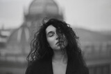 Fototapeta Zwierzęta - Young attractive brunette in a black jacket stands on the roof with closed eyes against the background of the urban space.