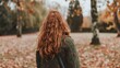 A young woman with long curly red hair wearing a green jacket enjoys a tranquil autumn day in a park, surrounded by fallen leaves