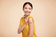 young asian women smiling after getting a vaccine, holding down her shirt sleeve and showing her arm with bandage after receiving vaccination on beige background,