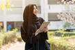 Closeup and crop cheerful Muslim student wearing traditional clothes stand and holding a tablet on walkway on her school building with sun flare background.