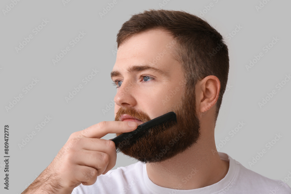 Young man brushing beard with comb on grey background, closeup