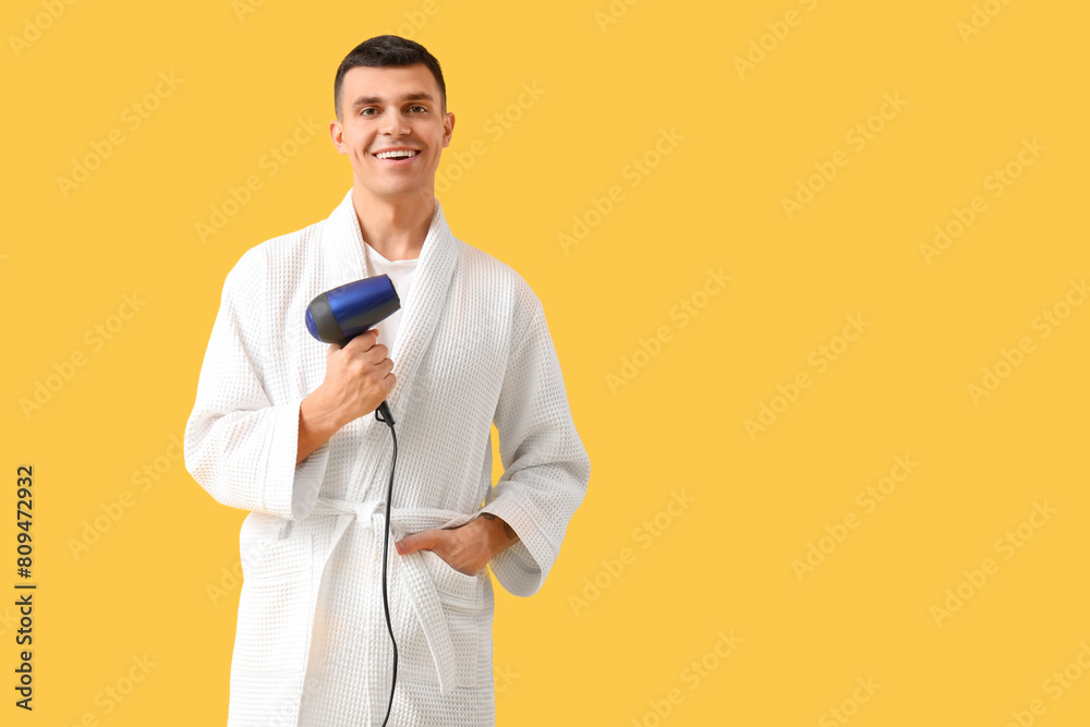Handsome young man in bathrobe with hair dryer on yellow background