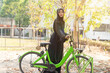 Cheerful Muslim student wearing traditional clothes stand and taking a bicycle in a public rental station in university with sun flare background.