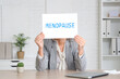 © Pixel-Shot - Mature businesswoman holding paper with word MENOPAUSE at table in office