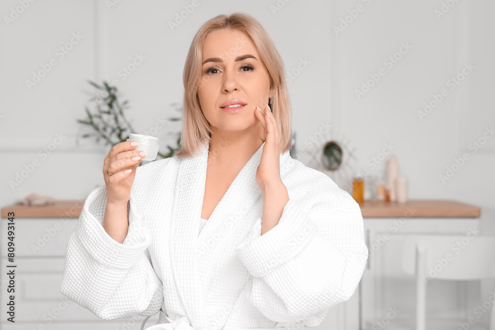 Beautiful mature woman with face cream taking care of her skin in bathroom