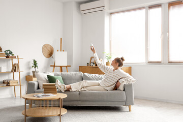 Wall Mural - Young woman turning on conditioner with remote control and lying on sofa in living room