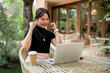 © bongkarn - A cheerful woman sits at a table, looking at her laptop with a happy face, celebrating good news.
