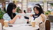 Two Asian women sit in a cafe at a mall, one excitedly showing off a new shirt to her friend.