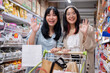 Two women smile and wave at the camera in a grocery store aisle, pushing a shopping cart together.