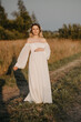 A young expectant mother in the third trimester of pregnancy in a white dress hugs her stomach against the backdrop of a natural landscape. The concept of future motherhood.