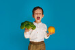A studio shot of a boy holding fresh broccoli and an orange on a blue background. The concept of healthy baby food.