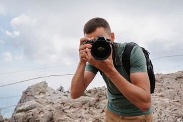 A young male tourist with a backpack climbed to the top of the mountain and takes pictures on the peak from a height. Facing the camera. The concept of active life and hiking.