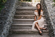 A young girl, a brunette with long hair, in a vintage dress is sitting on the steps of a gray stone staircase