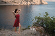 A young attractive Caucasian woman in a red dress at the edge of a cliff near the beach and the sea raised her hands up to the sun. The concept of freedom and travel