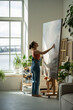 Calm artistic female adjusting fixing canvas with started, finished artwork. Thoughtful woman moving canvas with large big art painting, organizing workspace. Girl in sunny creative workshop studio.