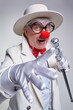 An elderly clown in a white robe and hat with a cane. The gloved hand is extended forward.