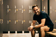 Portrait of a smiling fitness man sitting on the bench in the locker room and posing for the camera, dressed in sportswear.