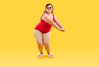 Fat lady in swimwear enjoying summer holiday and having fun at beach party. Full length portrait happy cheerful joyful young plus size woman wearing red swimsuit dancing isolated on yellow background
